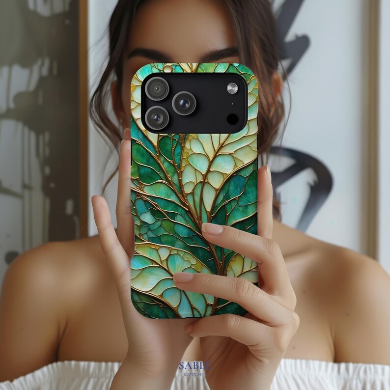May include: A smartphone case with a green and gold leaf design. The case features a detailed, stained-glass effect with shades of green, teal, and gold. The case has a black camera module and a gold-colored frame.
