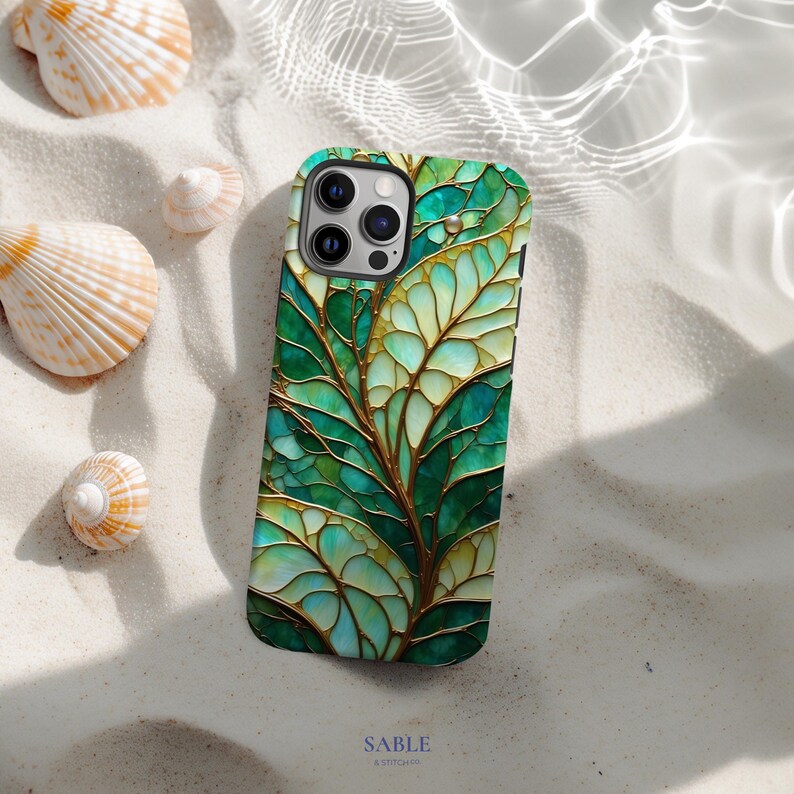 May include: A smartphone case with a green and gold leaf design. The case features a detailed, stained-glass effect with a network of veins. The case is on a sandy surface with seashells. The brand name "SABLE" is printed at the bottom.