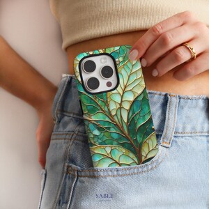 May include: A green and gold leaf design phone case. The case features a detailed pattern of leaves in shades of green and gold, with a black camera cutout. The phone case is in a denim pocket.