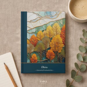 Pode incluir: Um diário de capa dura com lombada e capa azul-petróleo com um design colorido de árvores de outono. O diário é personalizado com o nome "Olivia" e o texto "A Journal of Gratitude & Joy". Um lápis e uma xícara de café estão no fundo.