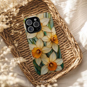 Op de afbeelding: Een telefoonhoesje met een bloemmotief van witte en gele narcissen met oranje centra. De hoes ligt op een geweven rieten dienblad. De merknaam "SABLE" is onderaan zichtbaar. De hoes heeft een glanzende afwerking.