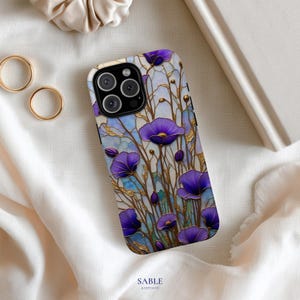 Può includere: Una custodia per telefono con un design a vetrata con fiori viola e dettagli dorati. La custodia ha un modulo fotocamera nero. Il marchio "SABLE & STITCH CO" è stampato in basso.