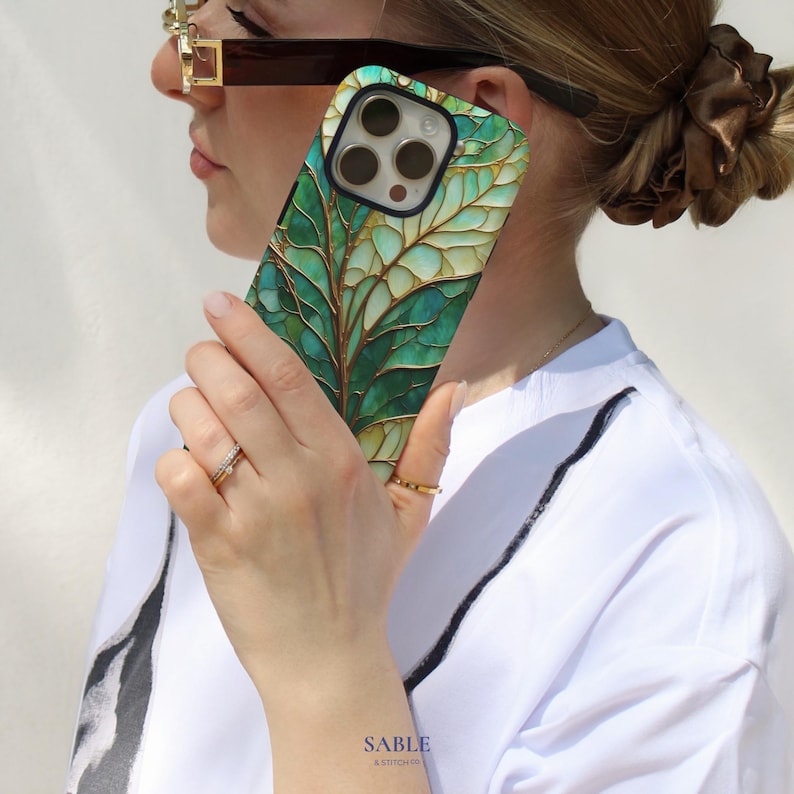 May include: A smartphone case with a green and gold leaf design. The case features a detailed illustration of leaves with gold accents, set against a teal and green background. The case has a black border and a camera cutout.