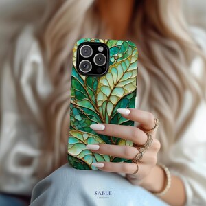 May include: Green and gold leaf pattern phone case. The case features a detailed design with shades of green and gold, resembling a stained-glass window. The phone case has a camera cutout and the text "SABLE & STITCH CO."