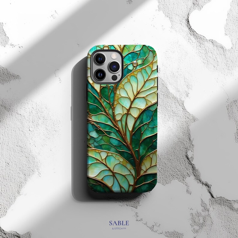 May include: A smartphone case with a leaf design. The case features a pattern of green and gold leaves with a textured appearance. The case has a glossy finish and is set against a white background. The brand name "SABLE & STITCH CO" is printed at the bottom.