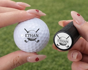 Könnte beinhalten: Ein weißer Golfball und ein schwarzer zylindrischer Stempel, beide mit einer schwarzen Grafik aus zwei gekreuzten Golfschlägern hinter einem Golfball und dem Namen ETHAN in der Mitte. Der Stempel wird gehalten, um das Motiv zu zeigen.