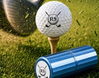 Könnte beinhalten: Ein weißer Golfball mit genoppter Oberfläche und einem schwarzen Logo mit den Buchstaben RS, gekreuzt von zwei Golfschlägern, auf einem Holz-Tee. Daneben steht ein zylindrisches blaues Metallgehäuse mit demselben RS-Logo auf der Endkappe.
