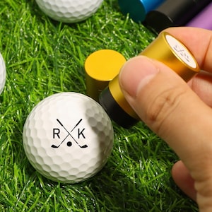 May include: White golf balls with the letters "R" and "K" and crossed golf clubs printed on them. A gold-colored stamp is being used to mark the golf balls. The golf balls are on a bed of green grass.