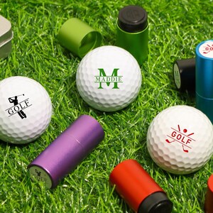 K&ouml;nnte beinhalten: Wei&szlig;e Golfb&auml;lle mit verschiedenen Aufdrucken und dem Wort "GOLF". Au&szlig;erdem sind kleine, farbige zylindrische Beh&auml;lter zu sehen. Die Artikel sind auf gr&uuml;nem Gras angeordnet. Einige Golfb&auml;lle haben eine Golfer-Grafik, andere das Wort "GOLF".