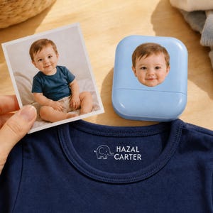 May include: A navy blue t-shirt featuring a white printed elephant icon and the text HAZAL CARTER on the inner neckline, accompanied by a light blue square stamp device displaying a child's face and a printed photograph of the same child.
