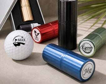 Könnte beinhalten: Ein weißer Golfball mit einer schwarzen Flagge und dem Text MAX, neben mehreren zylindrischen Metallbehältern in Schwarz, Blau, Rot und Grün. Ein Behälter zeigt den Text MR, ein anderer KIEVEN und ein weiterer eine Golfballgrafik mit dem Text MAX.