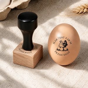 May include: A wooden rubber stamp with a black handle and a square base, featuring the text "HARVEST FARM FRESH EGGS" and a chicken illustration. Next to it is a brown egg stamped with the same "HARVEST FARM FRESH EGGS" design and chicken.