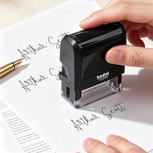 May include: A black Trodat Printy 4913 self-inking rubber stamp with a clear base, positioned on white paper. The stamp features the brand name and model number printed on the front. The paper displays the name Arthur Scott in black cursive ink.