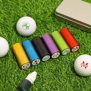 K&ouml;nnte beinhalten: Eine Auswahl an Golfzubeh&ouml;r, darunter personalisierte Golfb&auml;lle und zylindrische Beh&auml;lter. Die Golfb&auml;lle sind wei&szlig; mit individuellen gr&uuml;nen und roten Initialen. Die Beh&auml;lter sind in verschiedenen Farben wie Gelb, Blau, Schwarz, Lila, Gr&uuml;n und Rot.