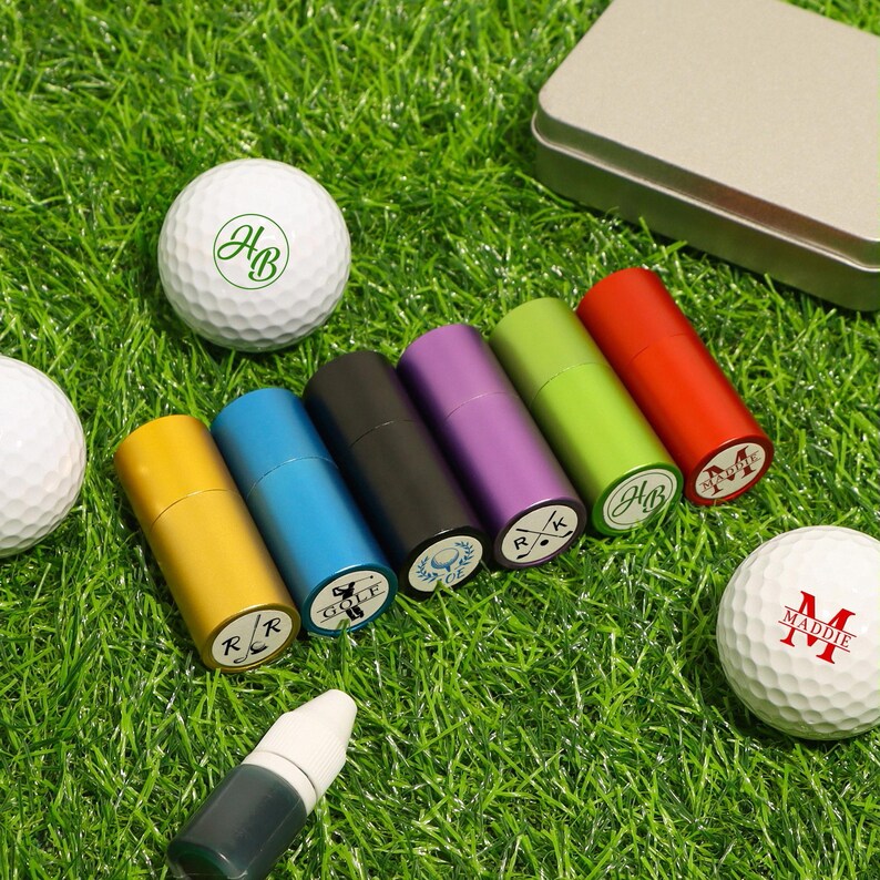 K&ouml;nnte beinhalten: Eine Auswahl an Golfzubeh&ouml;r, darunter personalisierte Golfb&auml;lle und zylindrische Beh&auml;lter. Die Golfb&auml;lle sind wei&szlig; mit individuellen gr&uuml;nen und roten Initialen. Die Beh&auml;lter sind in verschiedenen Farben wie Gelb, Blau, Schwarz, Lila, Gr&uuml;n und Rot.