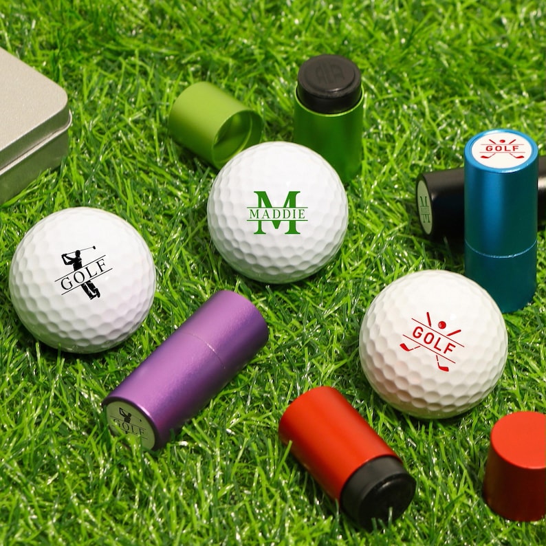K&ouml;nnte beinhalten: Wei&szlig;e Golfb&auml;lle mit verschiedenen Aufdrucken und dem Wort "GOLF". Au&szlig;erdem sind kleine, farbige zylindrische Beh&auml;lter zu sehen. Die Artikel sind auf gr&uuml;nem Gras angeordnet. Einige Golfb&auml;lle haben eine Golfer-Grafik, andere das Wort "GOLF".