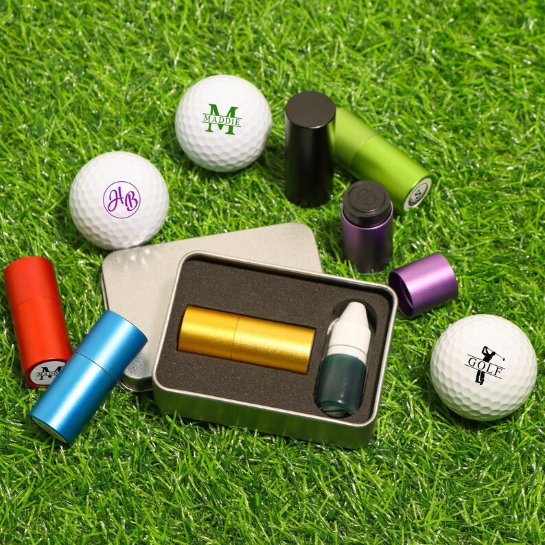 K&ouml;nnte beinhalten: Eine Sammlung von Golfzubeh&ouml;r, darunter personalisierte Golfb&auml;lle mit Initialen und Namen sowie verschiedene farbige Metallr&ouml;hren und Beh&auml;lter. Die Artikel sind auf gr&uuml;nem Kunstrasen angeordnet.