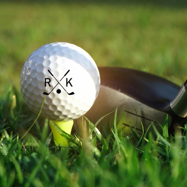 K&ouml;nnte beinhalten: Ein wei&szlig;er Golfball mit den Buchstaben "R" und "K" und gekreuzten Golfschl&auml;gern, der auf einem gelben Tee in gr&uuml;nem Gras liegt. Ein Golfschl&auml;ger befindet sich im Hintergrund und ist bereit, den Ball zu schlagen.