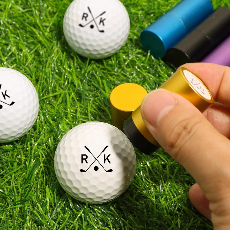 K&ouml;nnte beinhalten: Wei&szlig;e Golfb&auml;lle mit den Buchstaben "R" und "K" und gekreuzten Golfschl&auml;gern. Ein goldfarbener Stempel wird verwendet, um die Golfb&auml;lle zu markieren. Die Golfb&auml;lle liegen auf einer gr&uuml;nen Grasfl&auml;che.