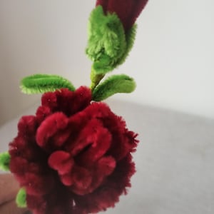 Puede incluir: Una flor roja hecha a mano con limpiapipas, hojas y tallo verdes. La flor está en plena floración, con un capullo al lado. La flor artesanal es una decoración única y caprichosa.