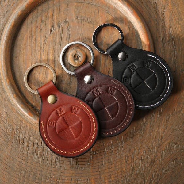 Puede incluir: Tres llaveros circulares de cuero en color tostado, marrón oscuro y negro, cada uno con una anilla de metal y el logotipo de BMW grabado. Las piezas de cuero están aseguradas con un remache metálico y tienen costuras visibles en el borde.