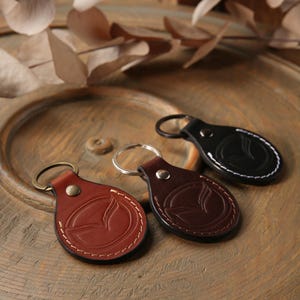May include: Three circular leather keychains featuring an embossed Mazda logo. The keychains are available in tan, dark brown, and black, each secured with a metal rivet and a circular split ring. Visible stitching runs along the perimeter.