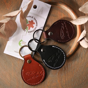 Op de afbeelding: Drie druppelvormige lederen sleutelhangers in donkerbruin, zwart en lichtbruin, elk met een metalen ring en een in reliëf gemaakt Jaguar-logo. Een wit productlabel met de tekst Sustainable Tanned Leather en Gold Rated is erachter zichtbaar.