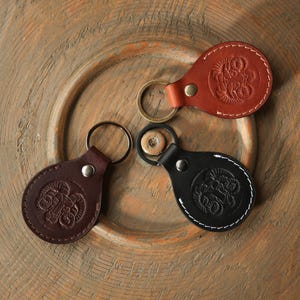 May include: Three leather keychains in brown, black, and red-brown, each with a metal keyring. The keychains have an oval shape with a decorative embossed design and are secured with a metal rivet. The keychains are on a wooden surface.