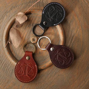 Könnte beinhalten: Drei Leder-Schlüsselanhänger in Schwarz, Braun und Rotbraun, jeweils mit rundem Design und silbernem Schlüsselring. Die Schlüsselanhänger zeigen ein geprägtes Waage-Symbol und weiße Nähte an den Rändern.
