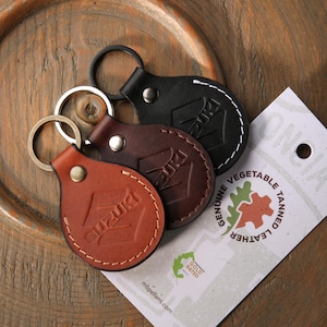 May include: Three leather keyrings in shades of brown and black, each with the "SUZUKI" logo embossed. The keyrings have metal rings and are stitched with contrasting thread. A tag with "GENUINE VEGETABLE TANNED LEATHER" is visible.