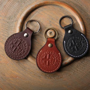 May include: Three leather keyrings in shades of brown and black, each featuring a circular emblem and the text "ALFA ROMEO MILANO." The keyrings have metal rings and are displayed on a wooden surface.