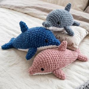 Könnte beinhalten: Drei handgefertigte Häkeltier-Delfin-Plüschtiere in Blau- und Rosatönen. Die Delfine haben schwarze Knopfaugen und weiße Bäuche. Sie liegen auf einem Bett mit neutralfarbener Bettwäsche.