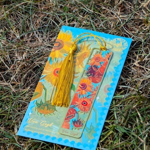 Puede incluir: Un conjunto de marcap&aacute;ginas y tarjetas con un dise&ntilde;o de girasoles inspirado en Van Gogh. El marcap&aacute;ginas tiene una borla dorada. La tarjeta tiene el texto "Van Gogh" y una cita. La paleta de colores incluye amarillo, naranja y azul.