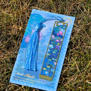 Puede incluir: Un marcap&aacute;ginas azul con un dise&ntilde;o de metal dorado de nen&uacute;fares, inspirado en Claude Monet. El marcap&aacute;ginas est&aacute; unido a una borla azul y descansa sobre una tarjeta azul con texto de la obra de Monet.