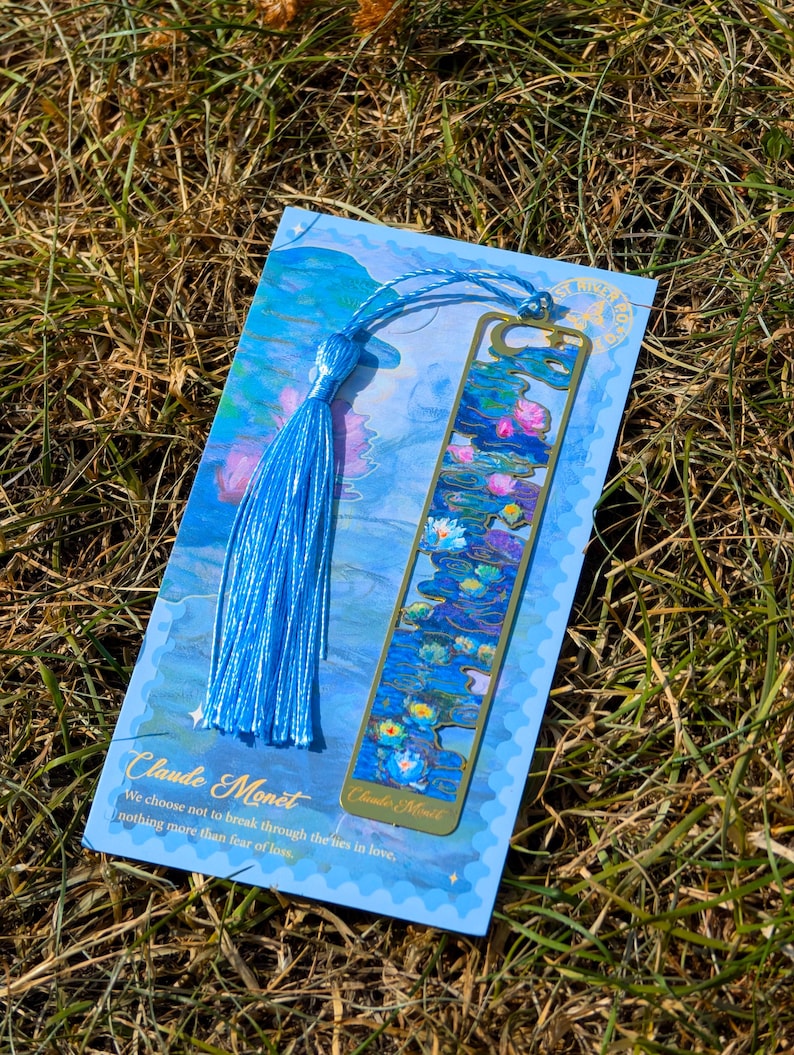 Puede incluir: Un marcap&aacute;ginas azul con un dise&ntilde;o de metal dorado de nen&uacute;fares, inspirado en Claude Monet. El marcap&aacute;ginas est&aacute; unido a una borla azul y descansa sobre una tarjeta azul con texto de la obra de Monet.