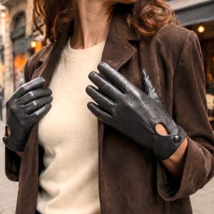Könnte beinhalten: Ein Paar schwarze Lederhandschuhe mit Perforationen an den Fingern und dem Handrücken sowie einem kleinen kreisförmigen Ausschnitt am Handgelenk, der mit einem einzelnen schwarzen Druckknopf verschlossen wird, für ein klassisches Design.