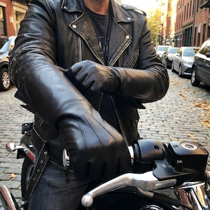 Può includere: Guanti e giacca da moto in pelle nera. I guanti sono lunghi, coprendo i polsi e parte degli avambracci. La giacca ha un classico stile biker con chiusura a zip e multiple tasche. La persona è seduta su una moto.