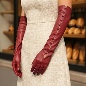 Peut inclure: Longs gants en cuir bordeaux avec des boutons. Les gants s'étendent au-delà des coudes et comportent une série de boutons sur le bras extérieur. Le design est classique et les gants sont en cuir lisse et souple.