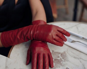 May include: A pair of long, vibrant red leather gloves. The gloves are resting on a marble-patterned table, with a dark-colored garment visible in the background. The gloves have a classic design, with individual fingers and a smooth texture.