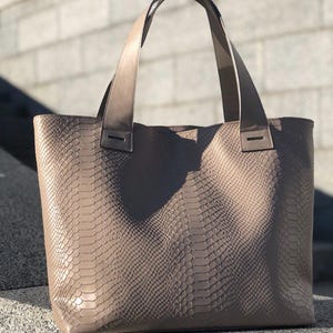 May include: A taupe tote bag with a snakeskin pattern. The bag has two long handles and a rectangular shape. The handles are attached to the bag with small, rectangular pieces of material. The bag is sitting on a stone surface.