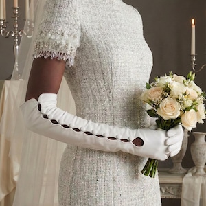 Può includere: Abito da sposa in tweed bianco con maniche corte, impreziosito da perline e paillettes. L'abito è abbinato a lunghi guanti bianchi con ritagli decorativi. Un bouquet di rose bianche e pesca è tenuto in mano.