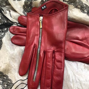 May include: A pair of red leather gloves featuring a vertical silver-toned metal zipper detail on the back of the hand. The interior of the glove displays a black label with the white text 6.5, indicating the size of the accessory.