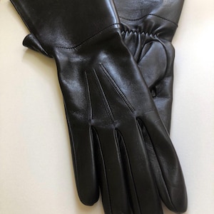 Puede incluir: Un par de guantes de cuero negro. Los guantes tienen un acabado liso y brillante y un diseño clásico. Los dedos están bien definidos y los puños son ligeramente alargados. Estos guantes son para el frío o para ocasiones formales.