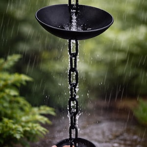 May include: Black rain chain with two round, bowl-shaped catchers connected by a chain. Water flows down the chain, creating a visual display. The background is a blurred green garden.