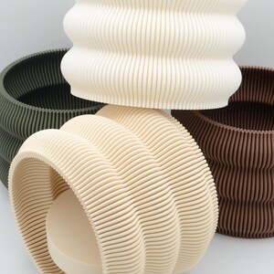 Könnte beinhalten: Eine Auswahl an gerippten Pflanzgefäßen in Creme, Beige, Olivgrün und Braun. Die Pflanzgefäße haben eine ausgeprägte, geriffelte Textur. Diese dekorativen Töpfe eignen sich für den Innen- und Außenbereich. Modernes und stilvolles Design.