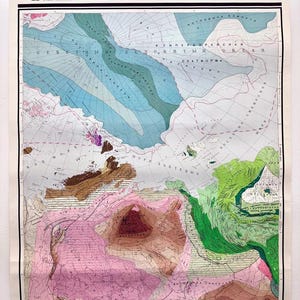 May include: A vintage map of Eurasia, featuring a detailed topographical representation with shades of blue, green, brown, and pink. The map is labeled in Cyrillic script and is held in place by colorful push pins. The map is mounted on a white surface.