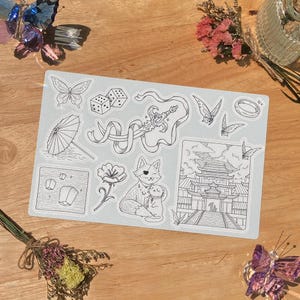 May include: A sheet of stickers with various black and white illustrations. Designs include butterflies, dice, a sword, a cat with a kitten, a pagoda, a flower, a parasol, a ring, and floating lanterns. The stickers are on a light blue background.
