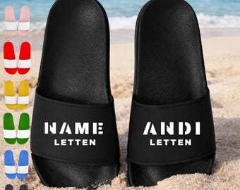 Puede incluir: Un par de sandalias negras tipo chancla con una banda ancha que muestra el texto blanco NAME LETTEN a la izquierda y ANDI LETTEN a la derecha. El texto "Designe deinen eigenen Style!" aparece arriba y hay pequeñas muestras de color a la izquierda.