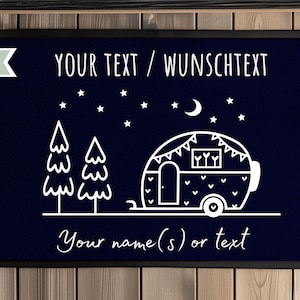 Puede incluir: Felpudo azul marino oscuro con borde negro. El felpudo presenta una ilustraci&oacute;n blanca de una caravana, &aacute;rboles, estrellas y una luna creciente. El texto incluye "YOUR TEXT / WUNSCHTEXT" y "Your name(s) or text".