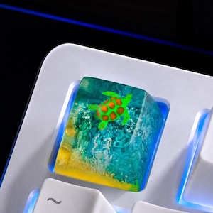 May include: A custom resin keycap featuring a miniature green sea turtle swimming in a translucent blue ocean scene with a yellow sandy bottom, installed on a white mechanical keyboard next to the F1 key and the number one key.